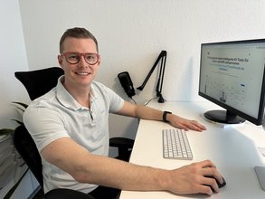 Man, der am Schreibtisch vor einem Bildschirm sitzt udn in die Kamera lächelt.