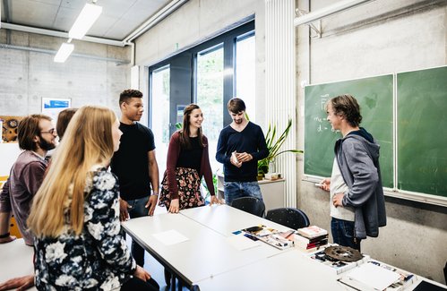 ©iStock Eine Gruppe junger Menschen steht mit einem Lehrer in einem modernen Klassenraum und unterhält sich lebhaft vor einer Tafel mit physikalischen Formeln.