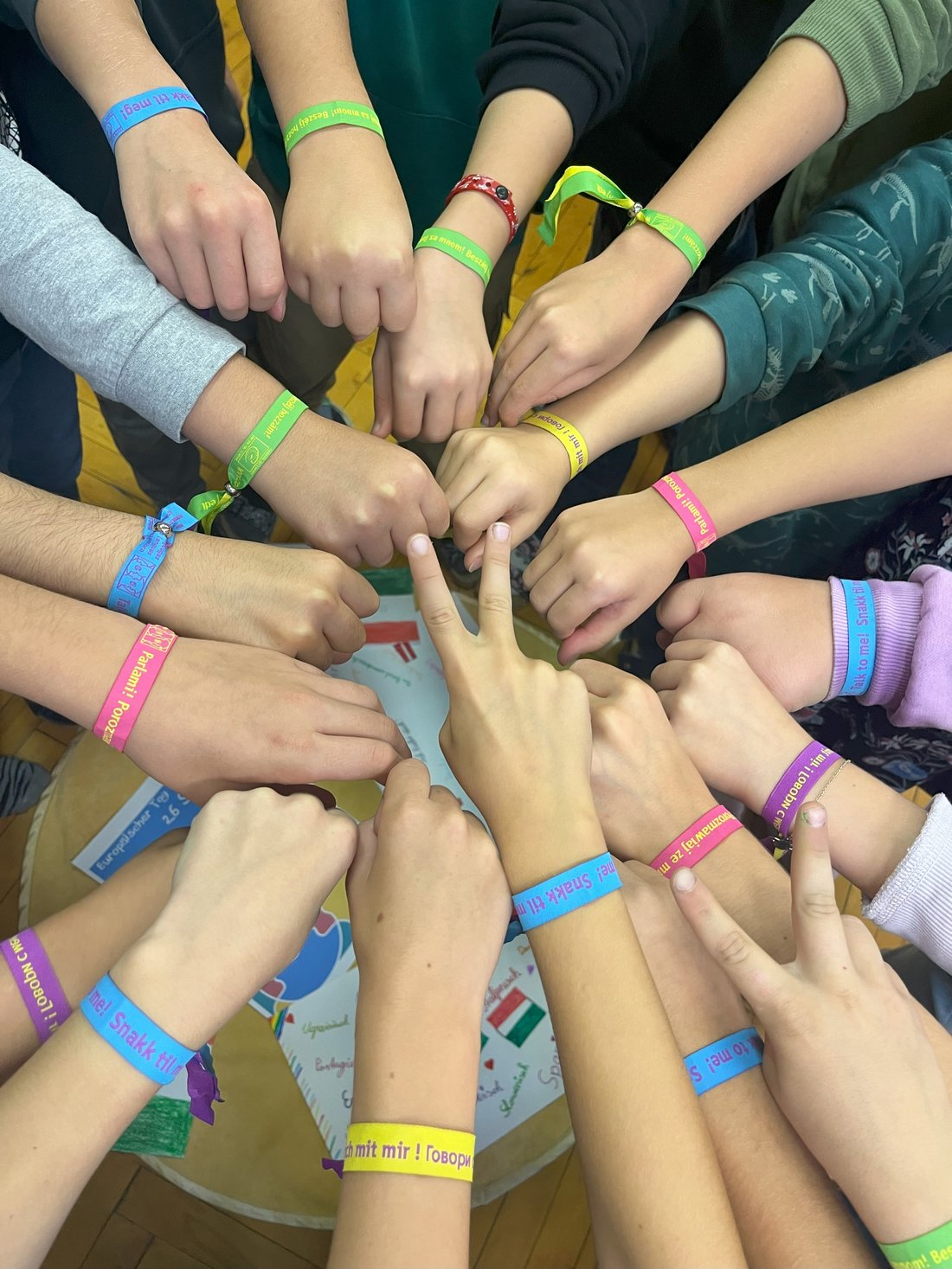 Arme mit bunten Armbändern. Zwei Hände zeigen zwei Finger wie ein "Peace"-Zeichen. Die anderen Hände sind zur lockeren Faust geballt. 