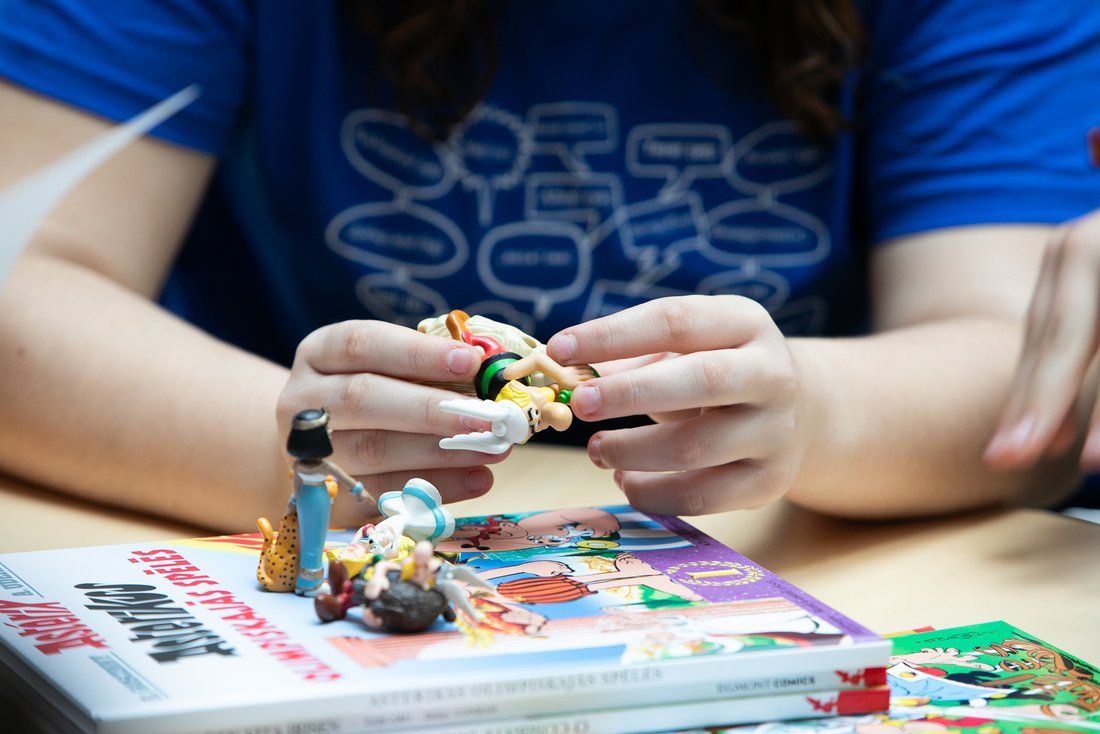 Nahaufnahme von zwei Händen, die kleine Asterix-Figuren halten. Die Figuren liegen auf einem Stapel von Asterix-Comics. Eine der Figuren scheint Obelix zu sein, und im Hintergrund ist eine Person in einem blauen T-Shirt mit einem Aufdruck von Sprechblasen zu sehen.
