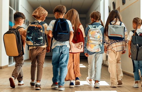 iStock Eine Gruppe von Grundschulkindern mit Rucksäcken läuft gemeinsam durch einen hellen Schulflur, von hinten aufgenommen.