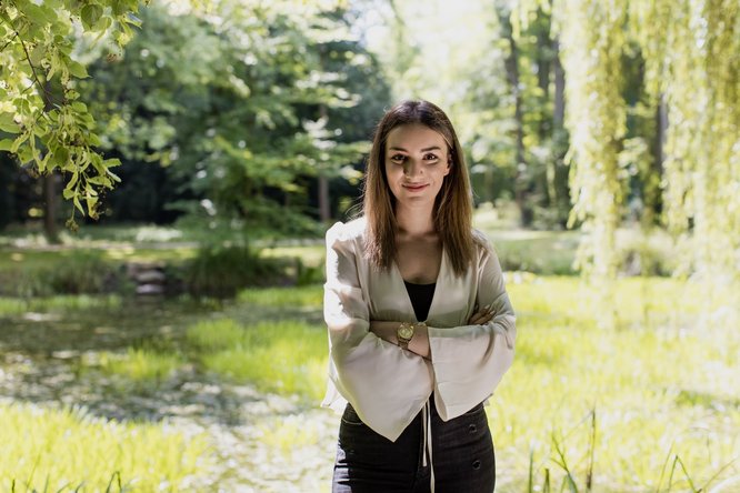 Das Bild zeigt die Interviewpartnerin, die lächelnd mit verschränkten Armen in einer grünen, natürlichen Umgebung steht. Sie trägt eine helle Bluse mit weiten Ärmeln und eine dunkle Hose. Im Hintergrund ist ein Teich und üppige Vegetation zu sehen, während Sonnenlicht durch die Bäume scheint und dem Bild eine freundliche, sommerliche Atmosphäre verleiht.