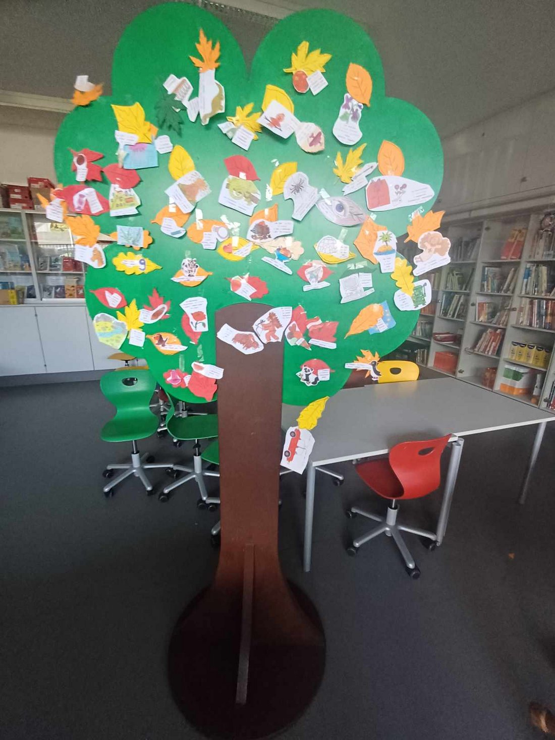 Baum aus Papier mit bunten Blättern und Textteilen darauf. Steht in einem Klassenzimmer mit einer Bücherwand rechts im Bild.