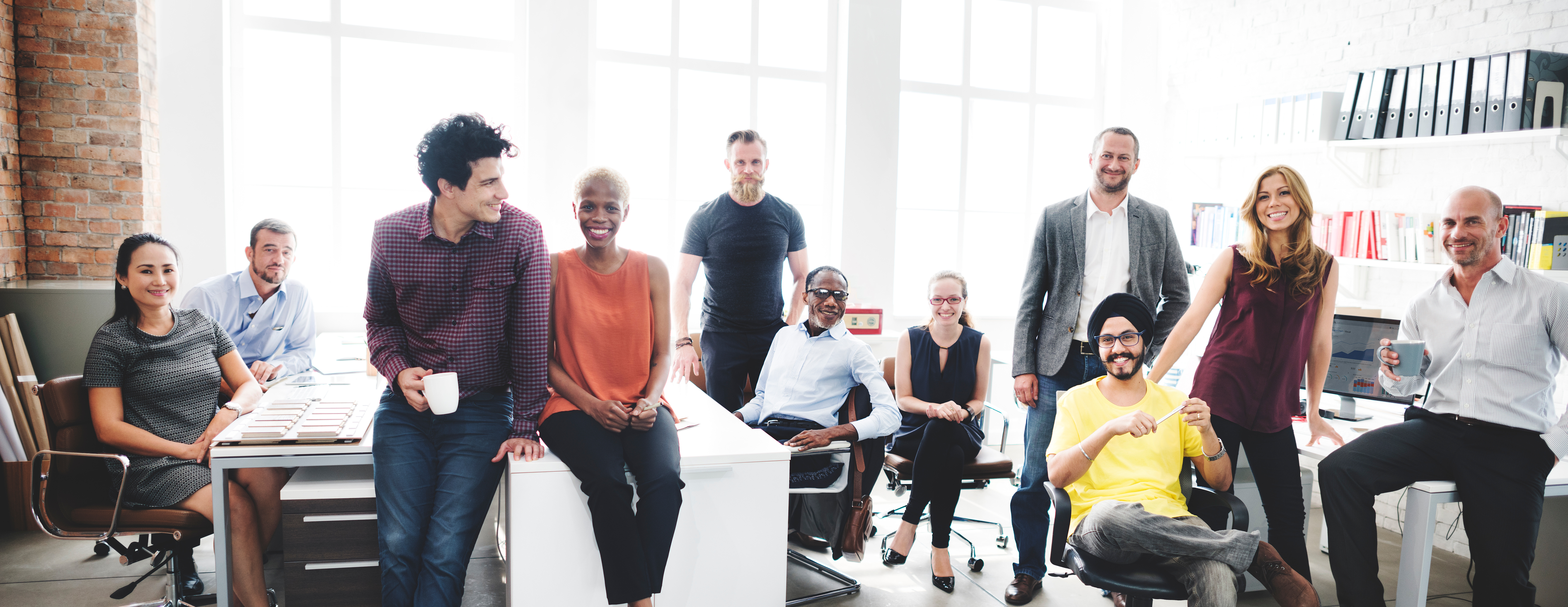 © Adobe Stock Eine Gruppe von zwölf Personen sitzt und steht in einem modernen Büro mit hellen, großen Fenstern. Sie wirken entspannt und lächelnd, während sie in die Kamera schauen. Einige halten Tassen in der Hand. Im Hintergrund sind Regale mit Ordnern und Büchern sowie Computerarbeitsplätze zu sehen. Die Szene vermittelt eine positive und kollegiale Arbeitsatmosphäre.