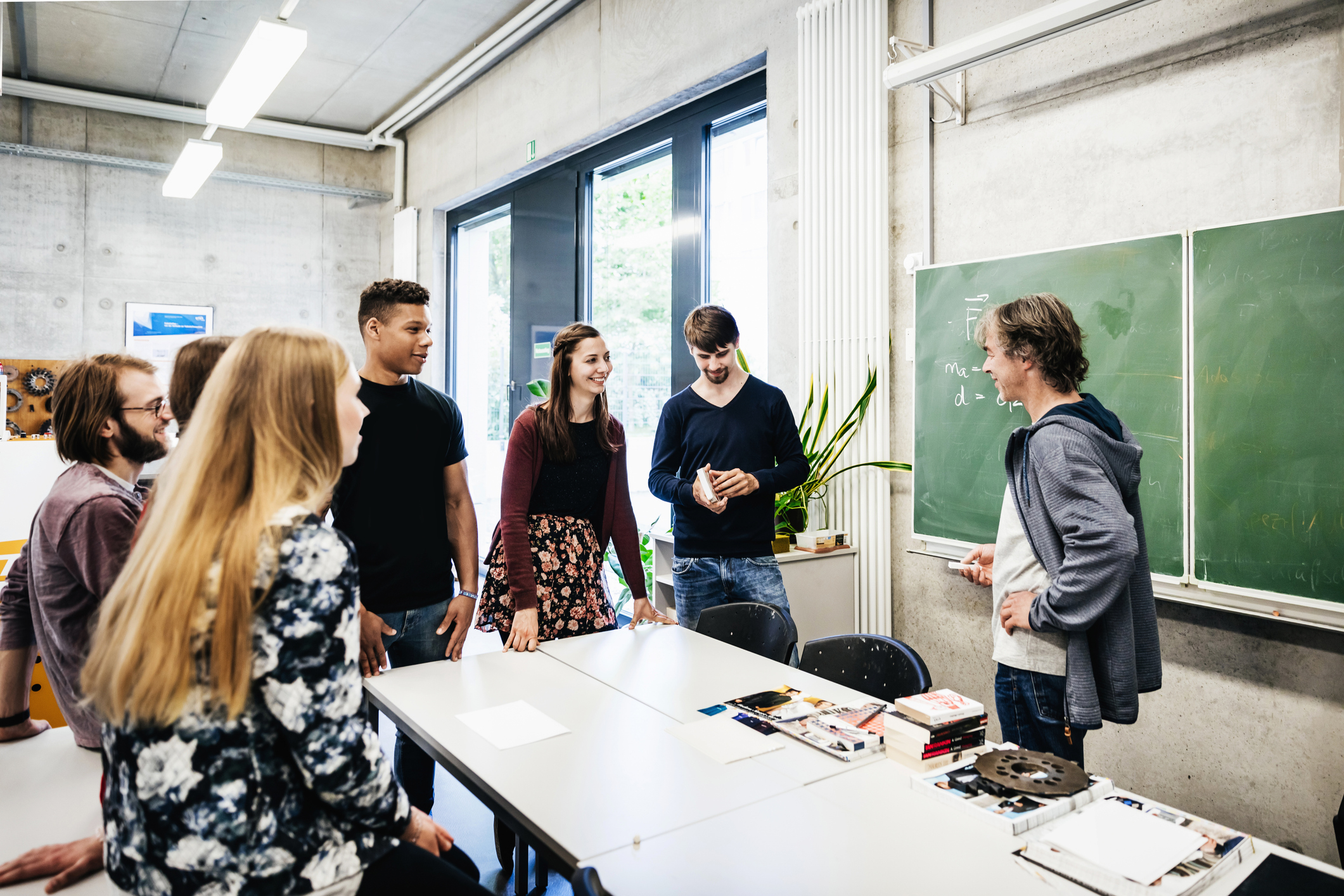 ©iStock Eine Gruppe junger Menschen steht mit einem Lehrer in einem modernen Klassenraum und unterhält sich lebhaft vor einer Tafel mit physikalischen Formeln.