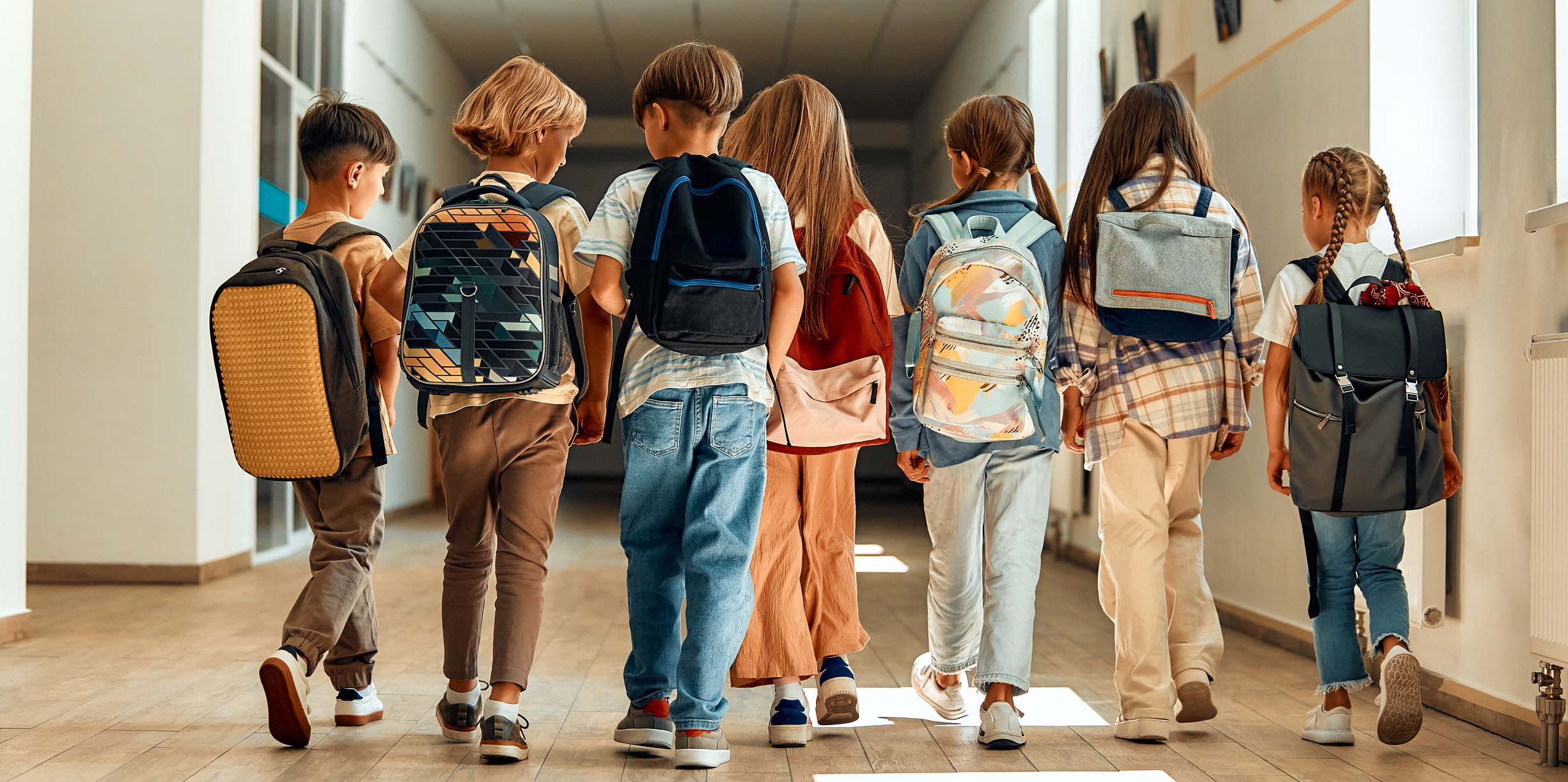 iStock Eine Gruppe von Grundschulkindern mit Rucksäcken läuft gemeinsam durch einen hellen Schulflur, von hinten aufgenommen.